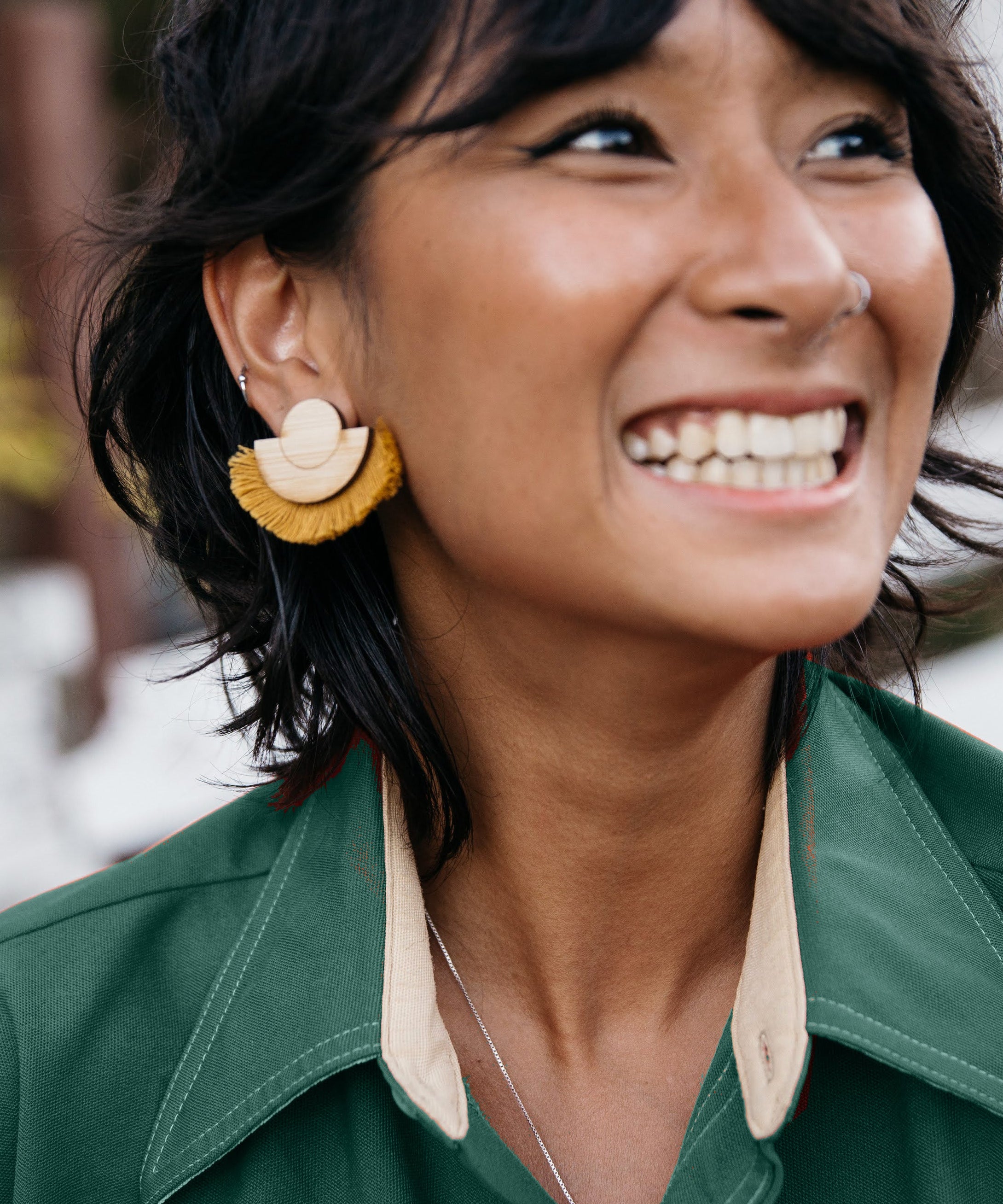 Woman wearing a mustard fringe and wood earrings and green shirt with a blurred natural background