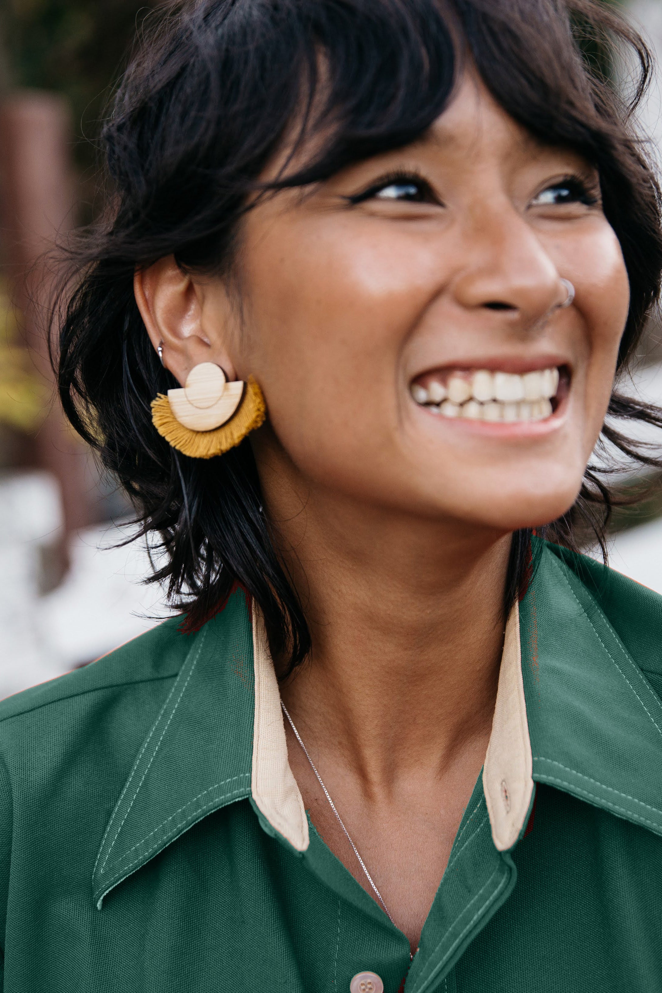 Woman wearing a mustard fringe and wood earrings and green shirt with a blurred natural background