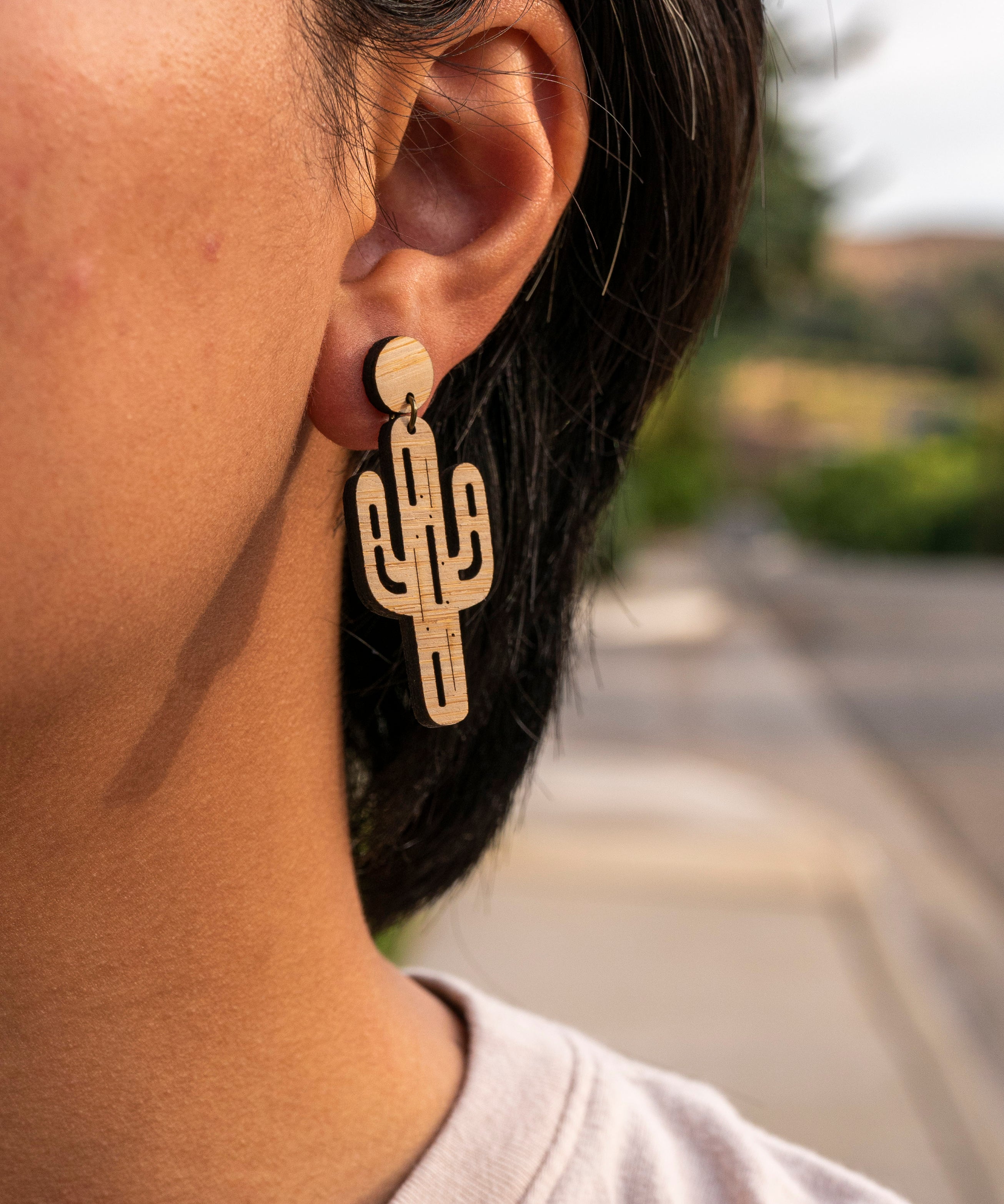 Laser cut bamboo cactus-shaped earrings worn by a person with a blurred outdoor background