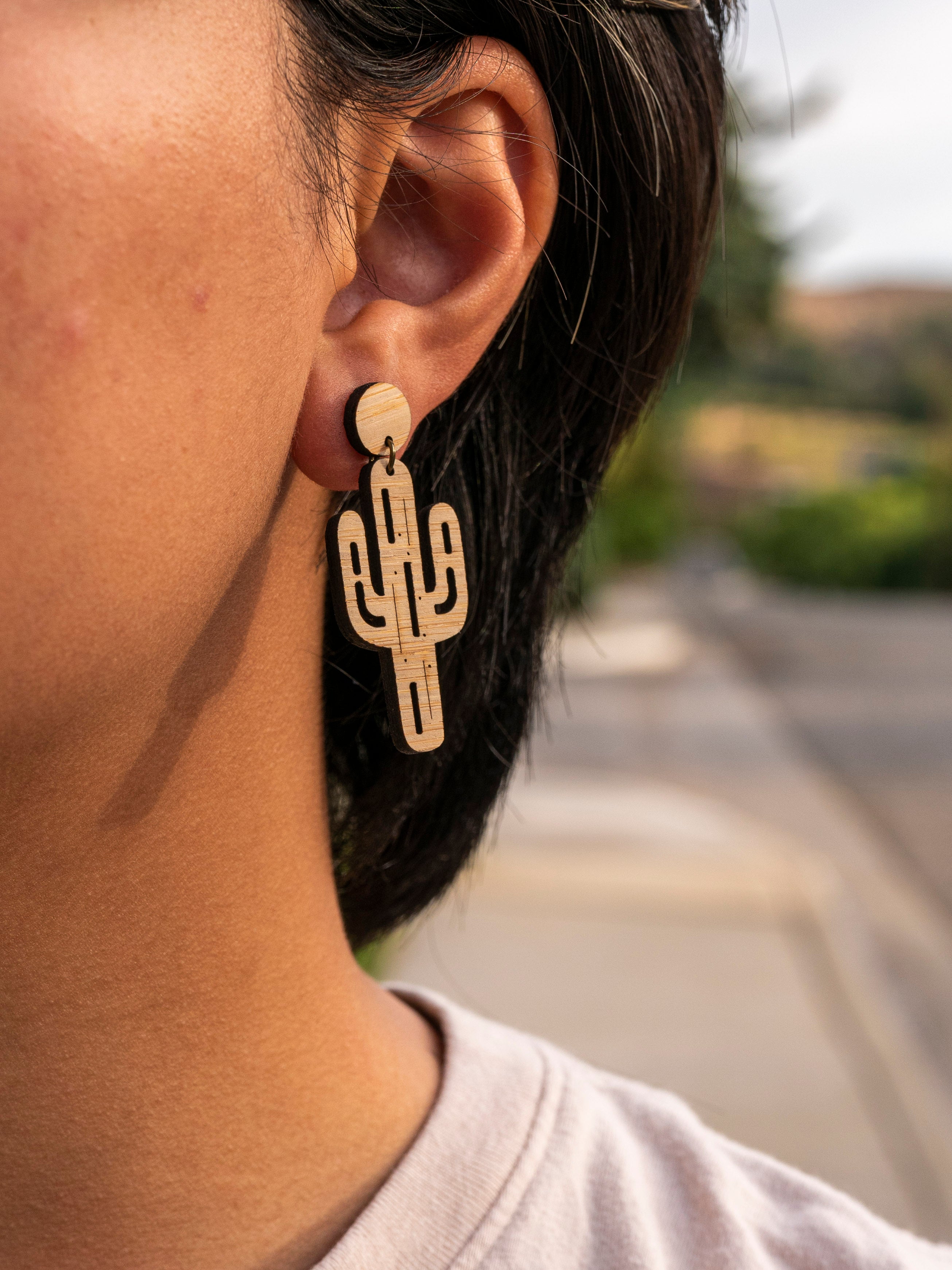Laser cut bamboo cactus-shaped earrings worn by a person with a blurred outdoor background