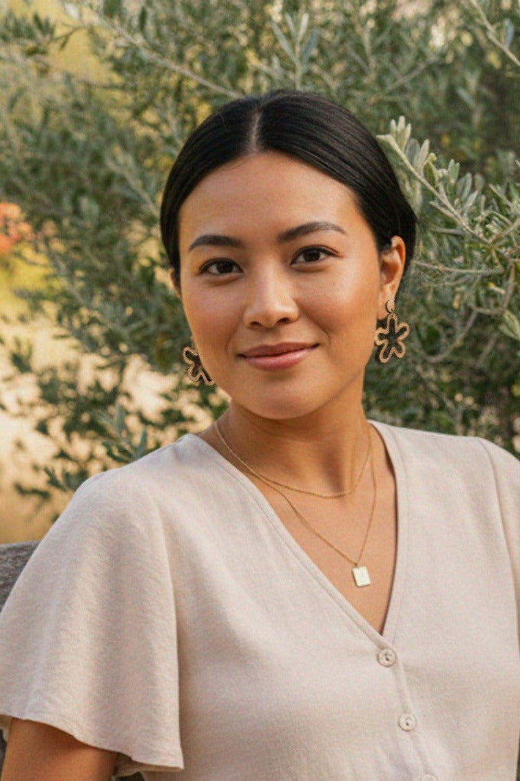 Woman wearing abstract flower cutout drop earrings, standing in front of green foliage.