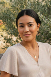Woman wearing abstract flower cutout drop earrings, standing in front of green foliage.