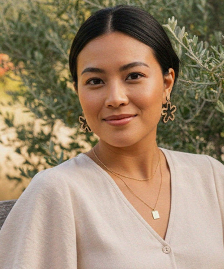 Woman wearing abstract flower cutout drop earrings, standing in front of green foliage.
