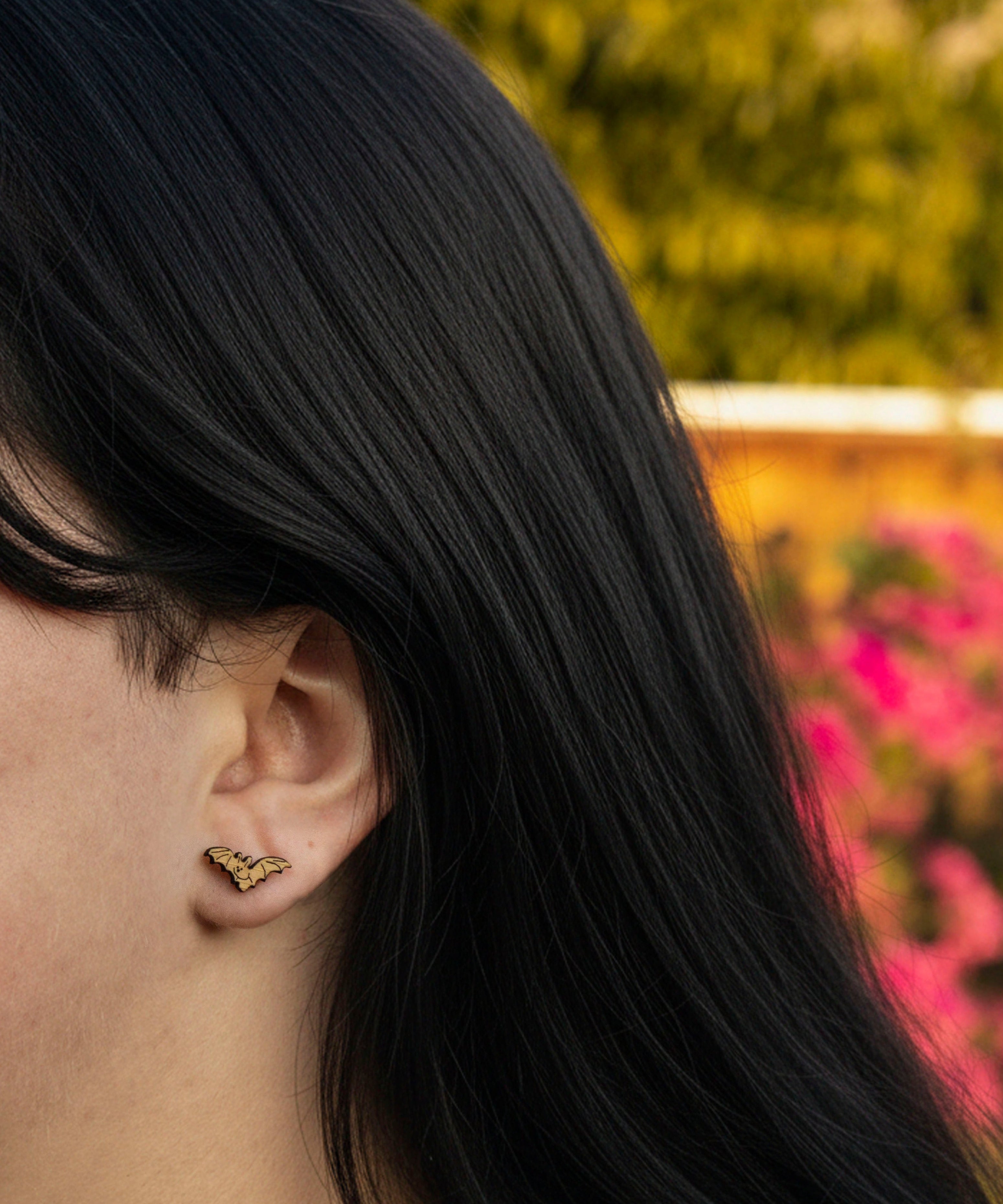 Close-up of a person with long dark hair wearing cute bat stud earrings, with a blurred natural background.