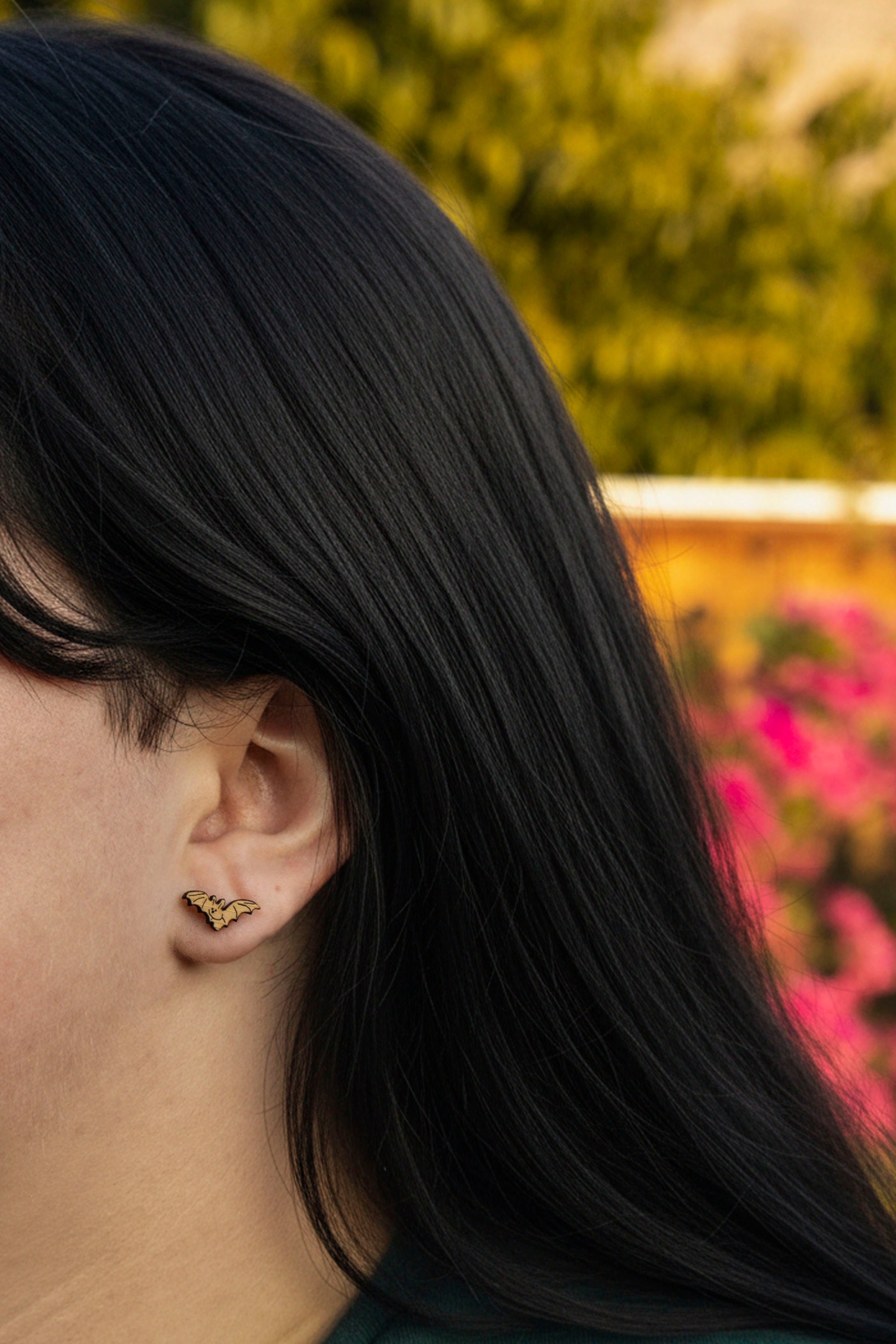 Close-up of a person with long dark hair wearing cute bat stud earrings, with a blurred natural background.