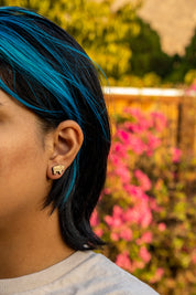 Person with blue hair wearing a bison wood earring in front of pink flowers and greenery