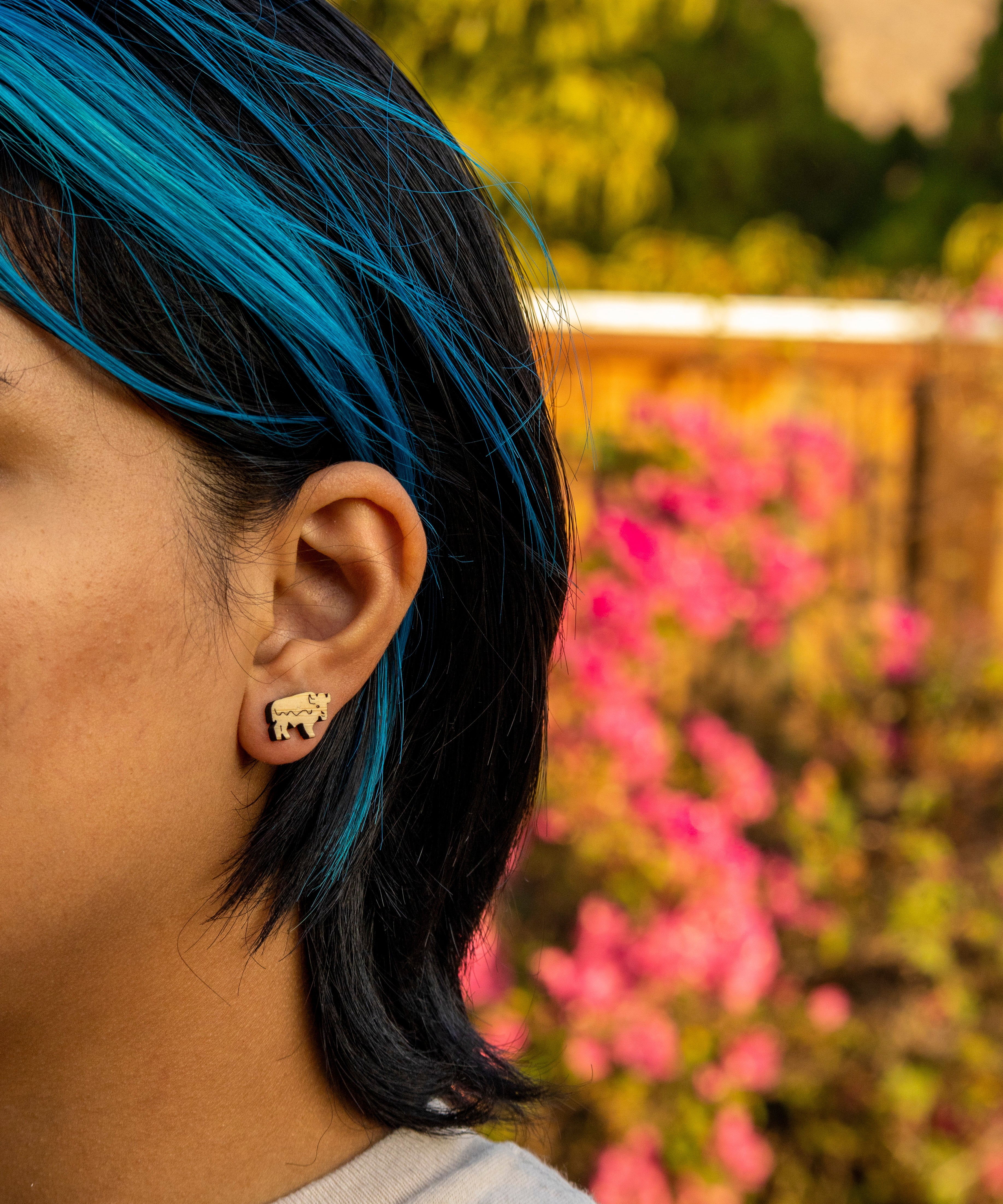 Person with blue hair wearing a bison wood earring in front of pink flowers and greenery
