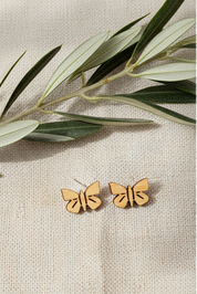 Wooden butterfly earrings on a beige fabric background with green leaves.