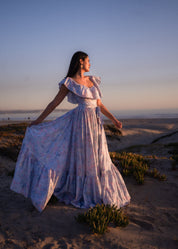 Woman wearing large bamboo daisy dangles earrings in a white floral dress standing on a sandy beach at sunset.
