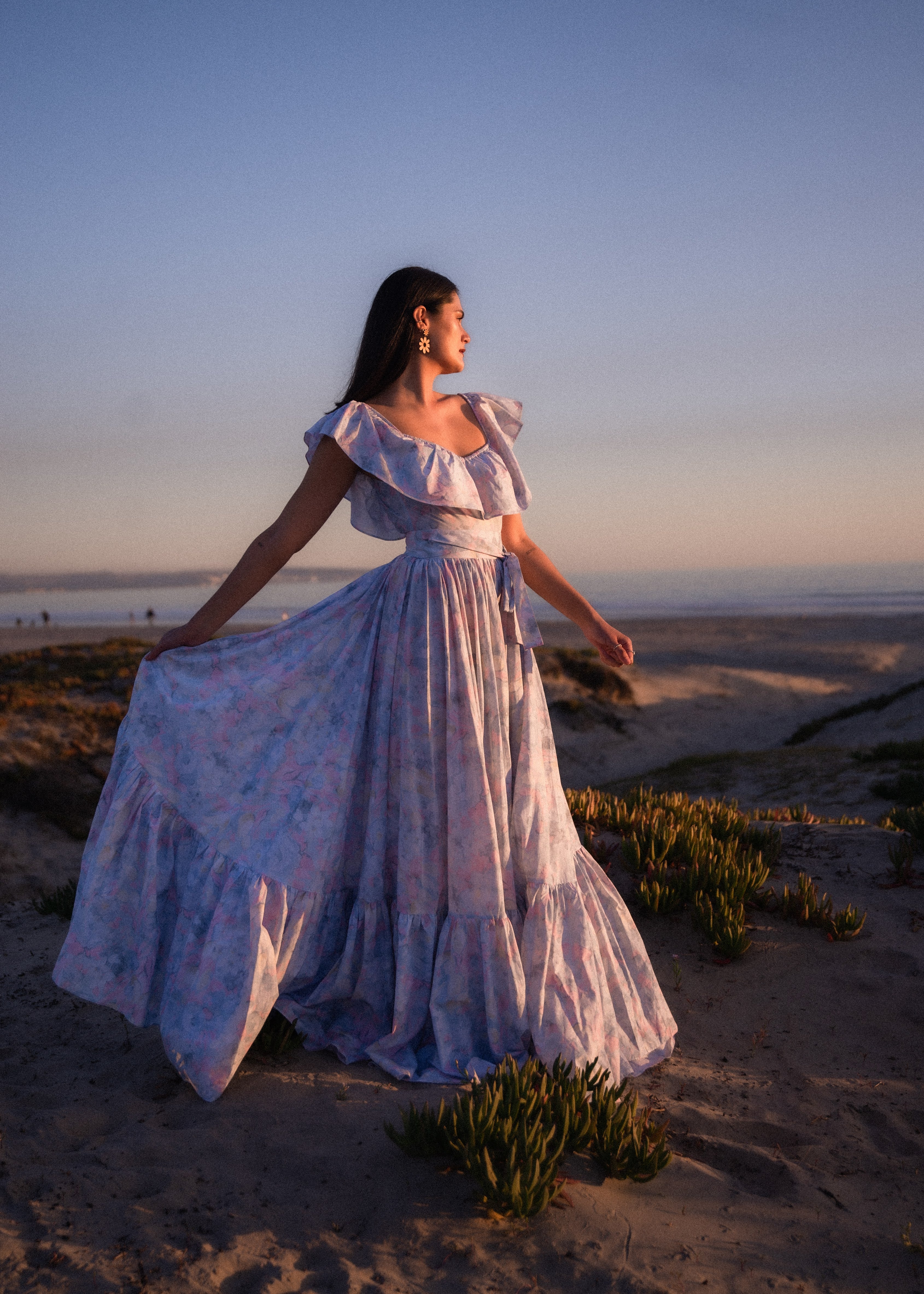 Woman wearing large bamboo daisy dangles earrings in a white floral dress standing on a sandy beach at sunset.