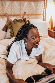 Woman wearing simple rainbow dangle wood earrings, laying on a bed in a cozy room.