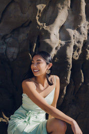 Woman wearing coral laser-cut bamboo earrings, in a light green dress sitting against a rocky wall on a beach