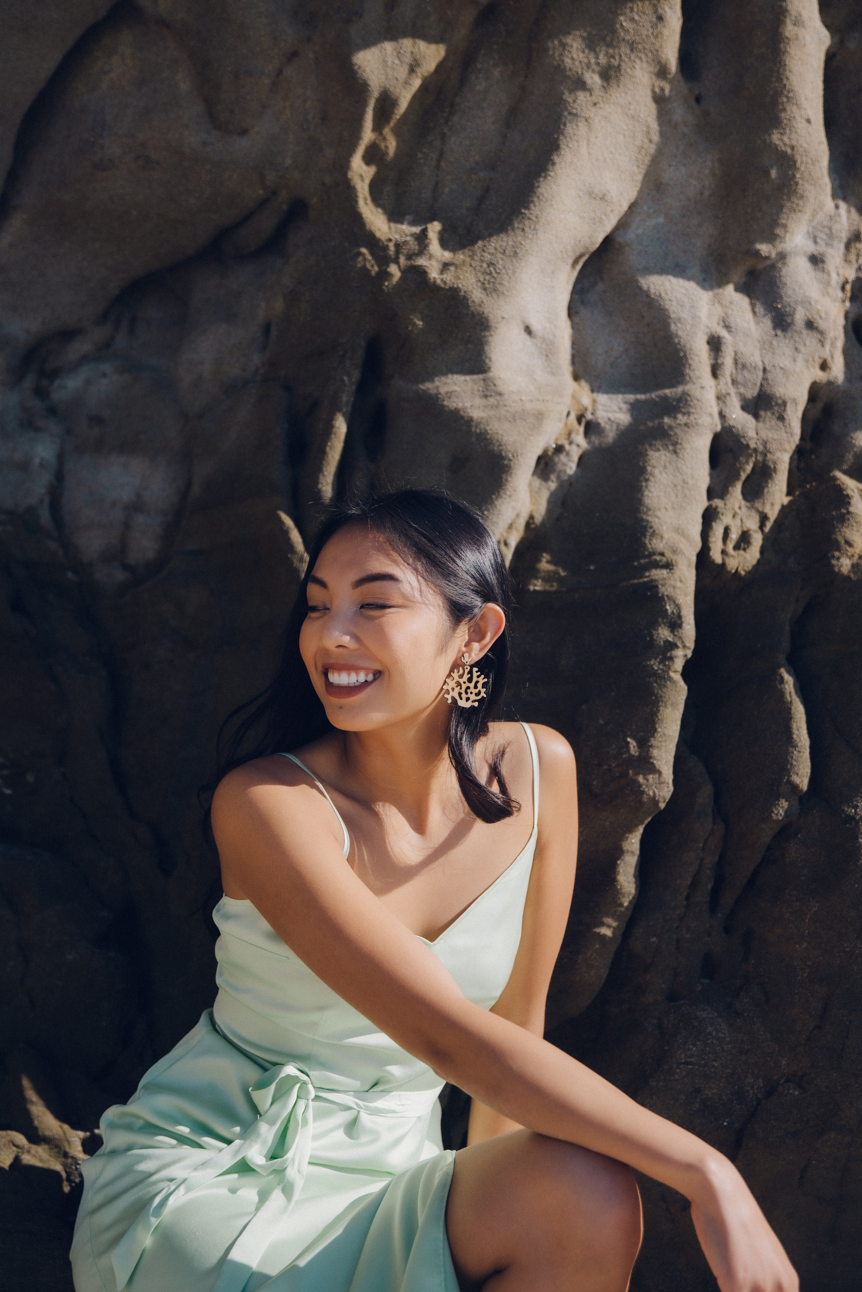 Woman wearing coral laser-cut bamboo earrings, in a light green dress sitting against a rocky wall on a beach