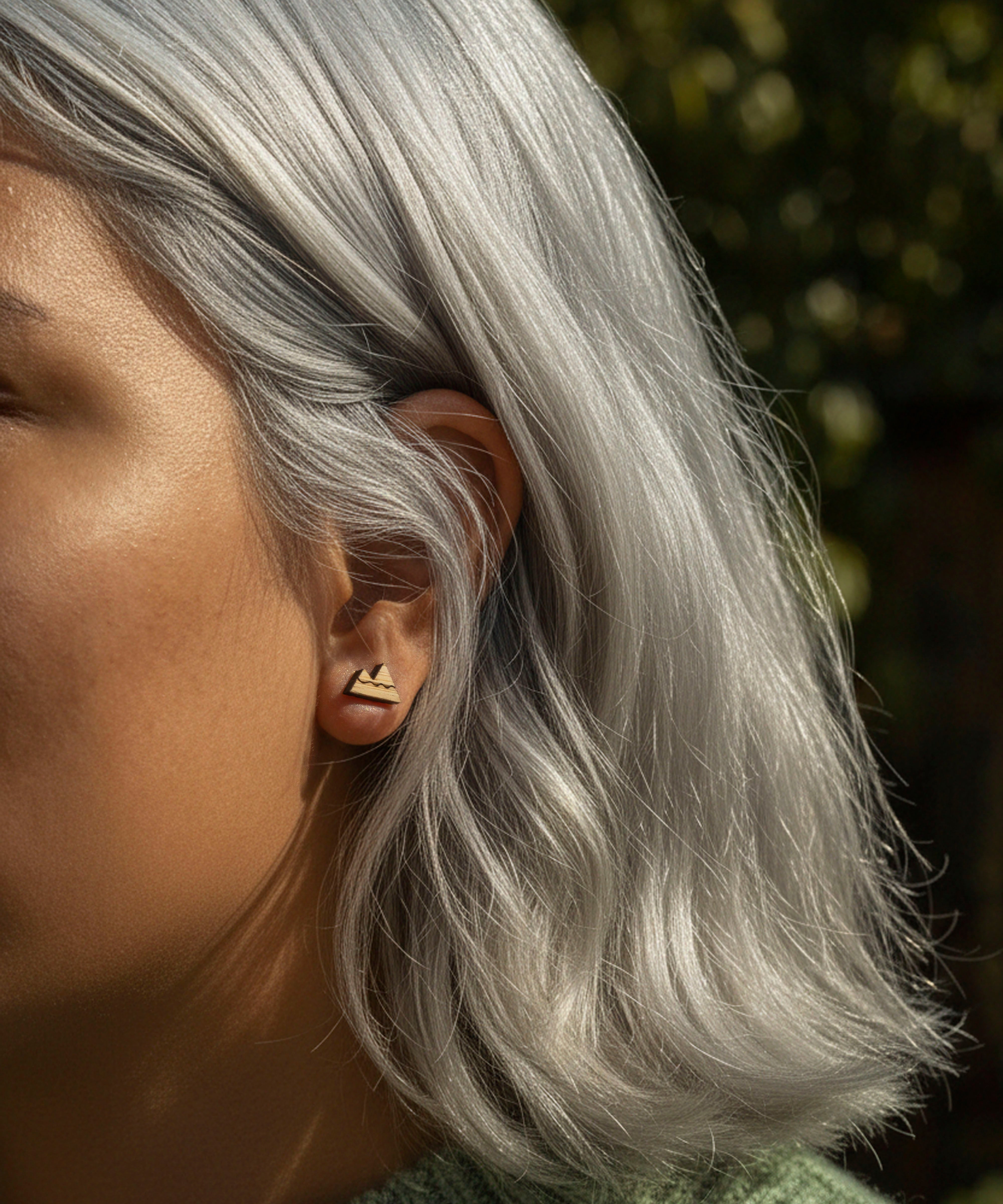 Close-up of a person with silver hair wearing mountain stud earring, with a blurred natural background.