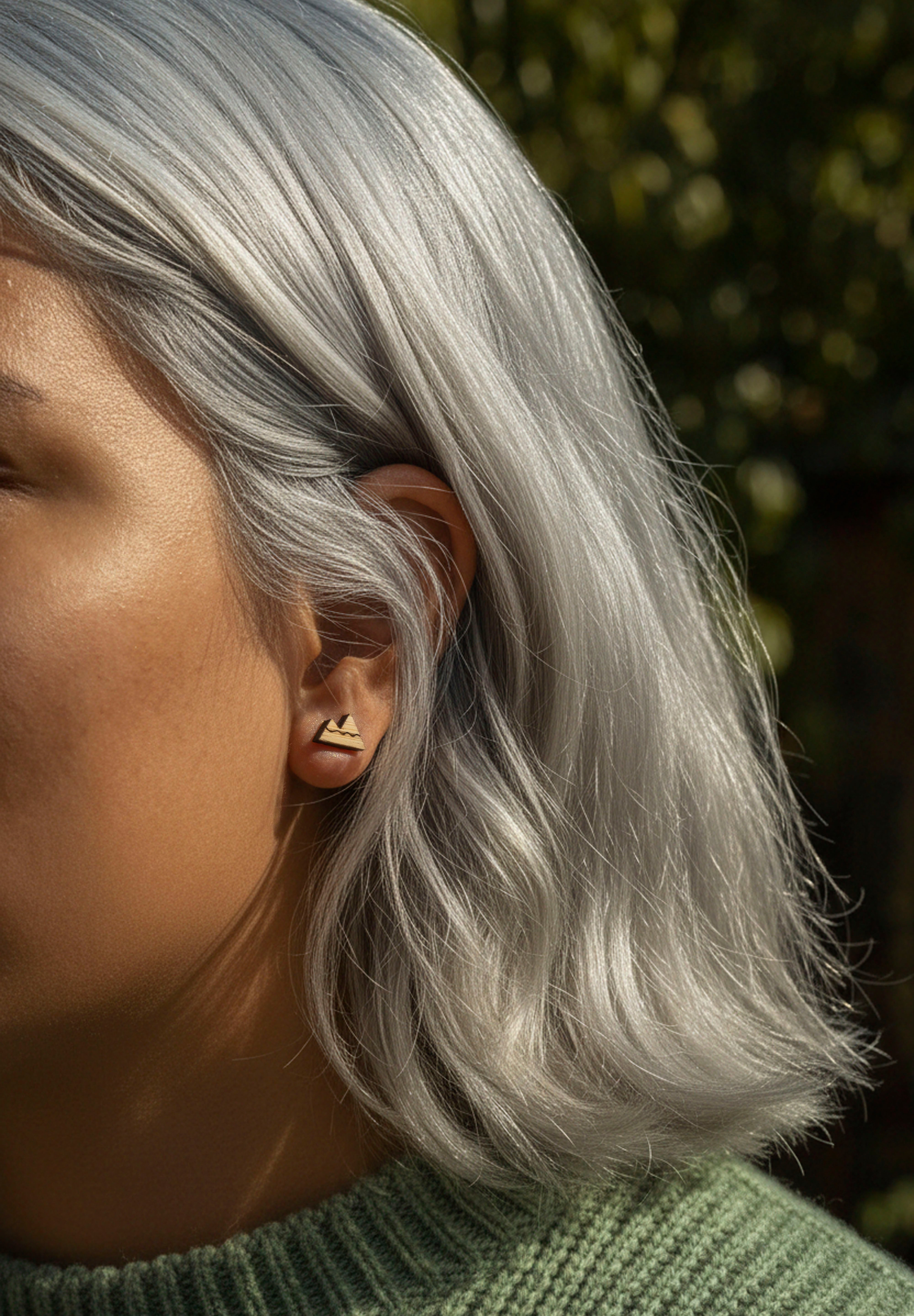 Close-up of a person with silver hair wearing mountain stud earring, with a blurred natural background.