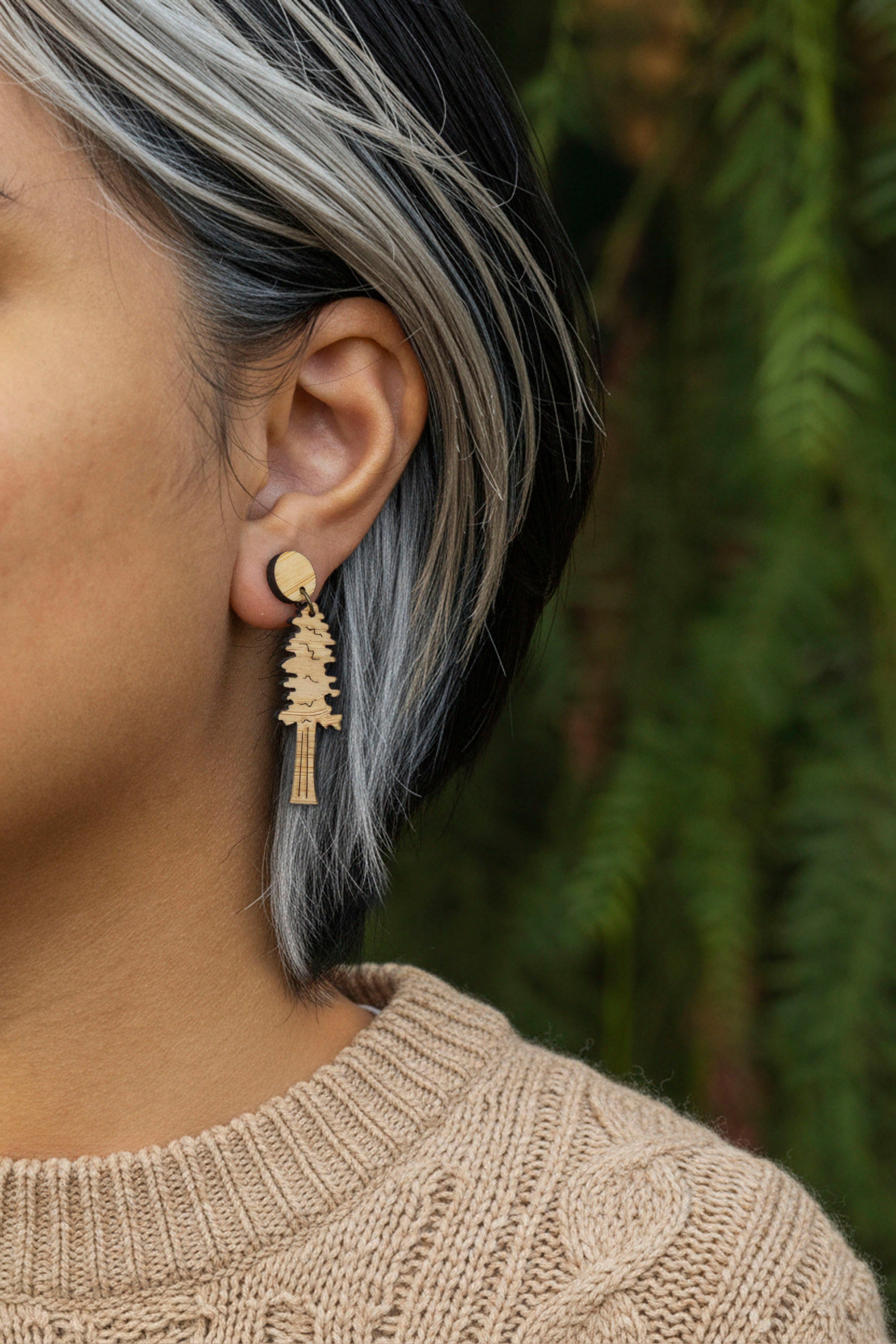 Close-up of a person wearing a wood earring with a pine tree design, against a blurred green background.