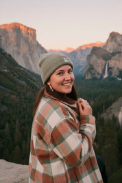Woman wearing redwood dangle earrings, in a plaid coat and beanie standing in front of Yosemite national park.