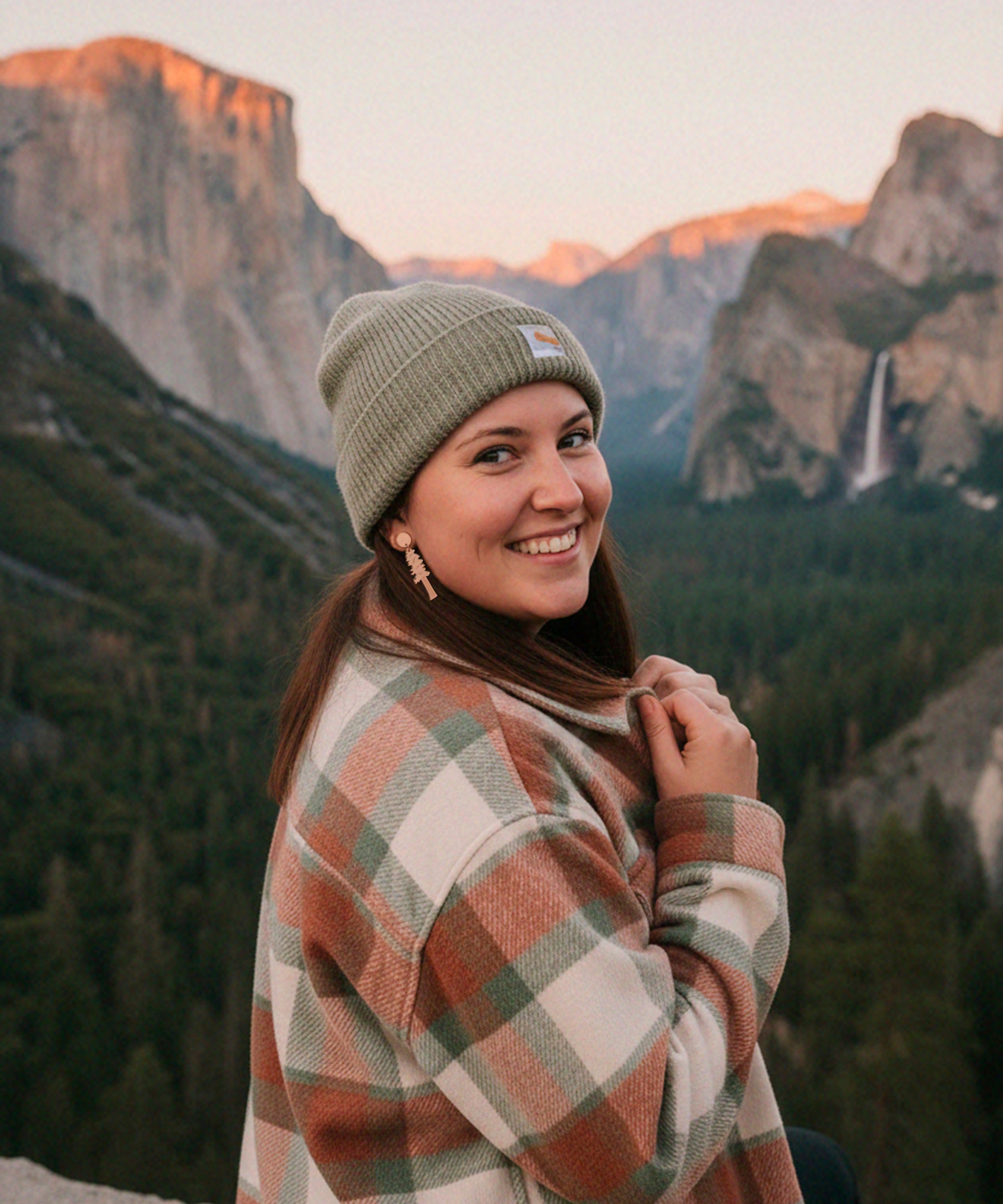 Woman wearing redwood dangle earrings, in a plaid coat and beanie standing in front of Yosemite national park.