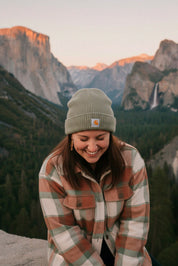 Person wearing redwood dangles earrings a plaid jacket and beanie with mountains in the background