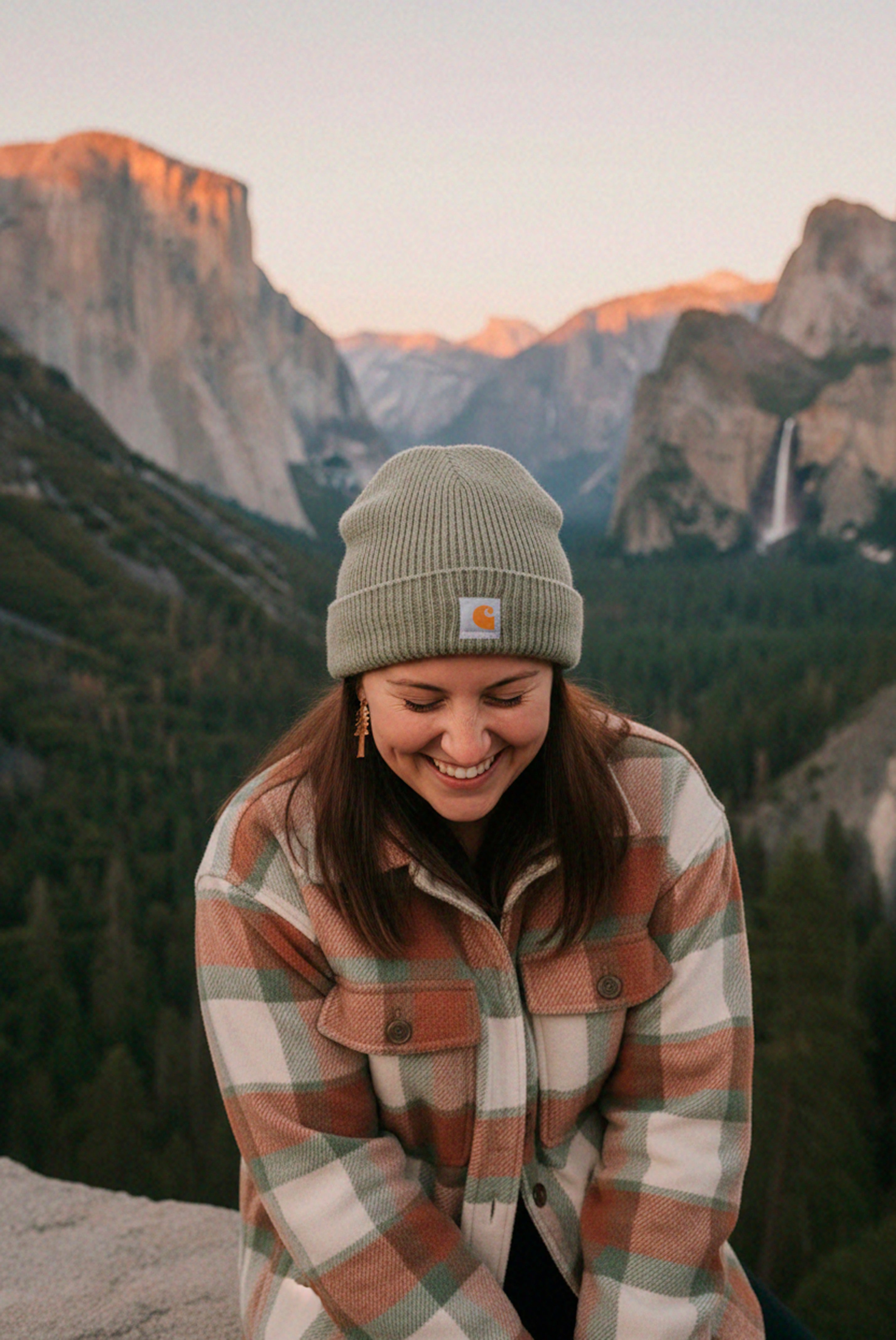 Person wearing redwood dangles earrings a plaid jacket and beanie with mountains in the background