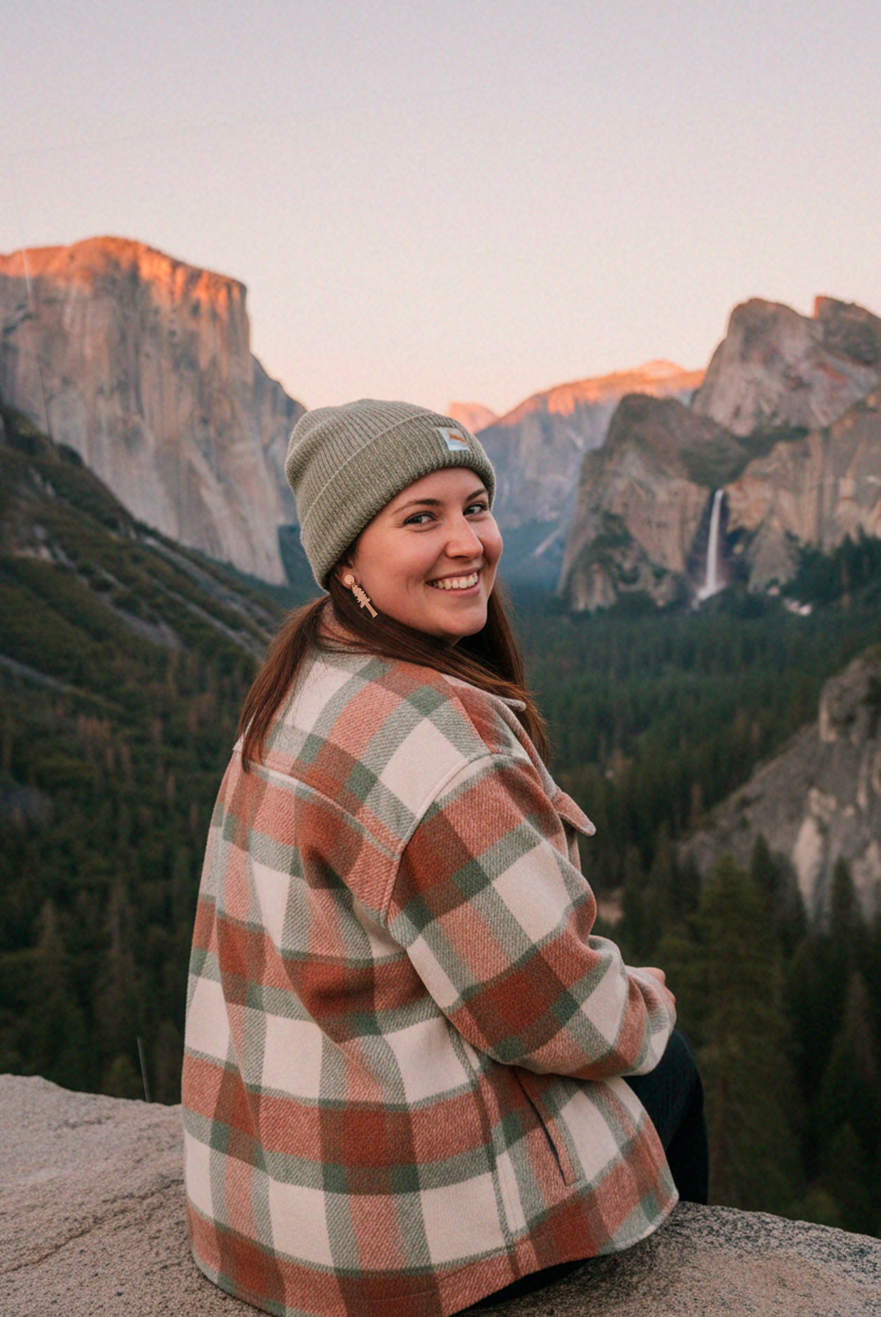 Person wearing a redwood dangle earrings, plaid shirt and beanie sitting on a rock with mountains in the background