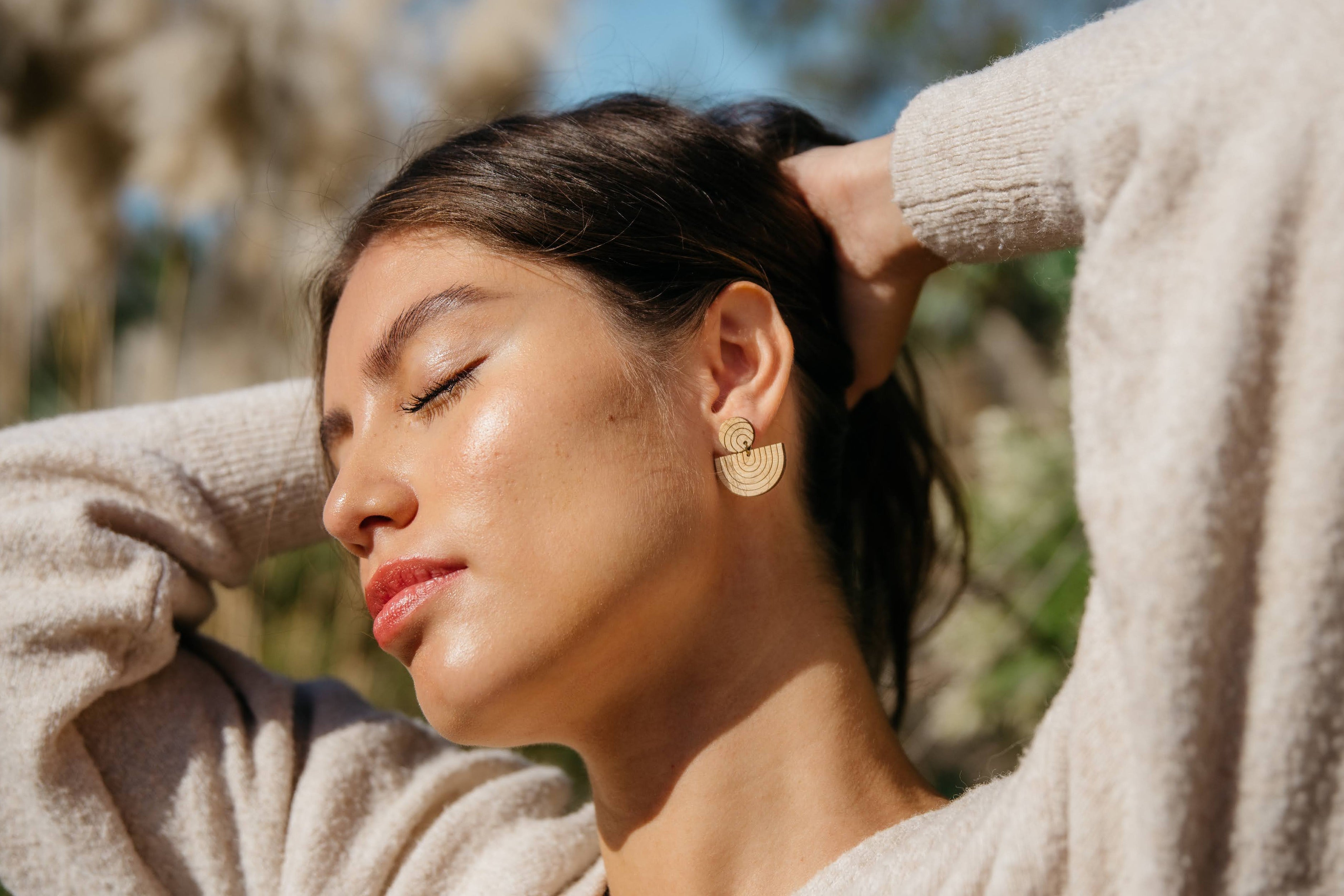 Woman wearing reverse rainbow dangle earrings in a beige sweater with eyes closed, standing outdoors with plants in the background