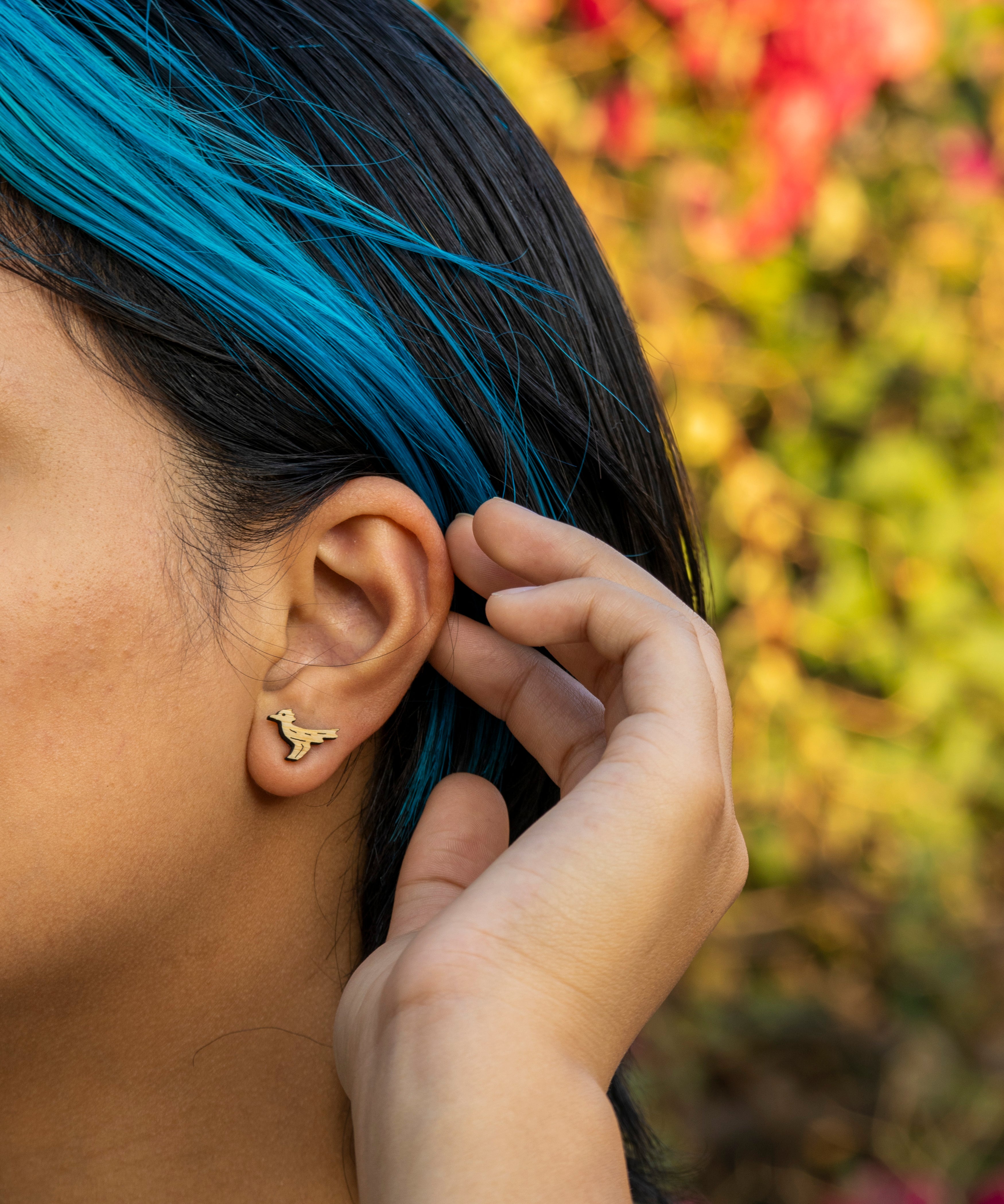 Person with blue hair adjusting a roadrunner wood earring outdoors