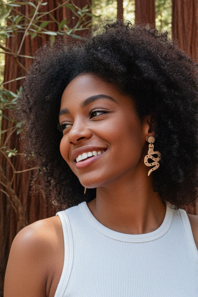 Woman with snake dangles earrings, and curly hair wearing a light blue tank top and gold earrings, standing outdoors.