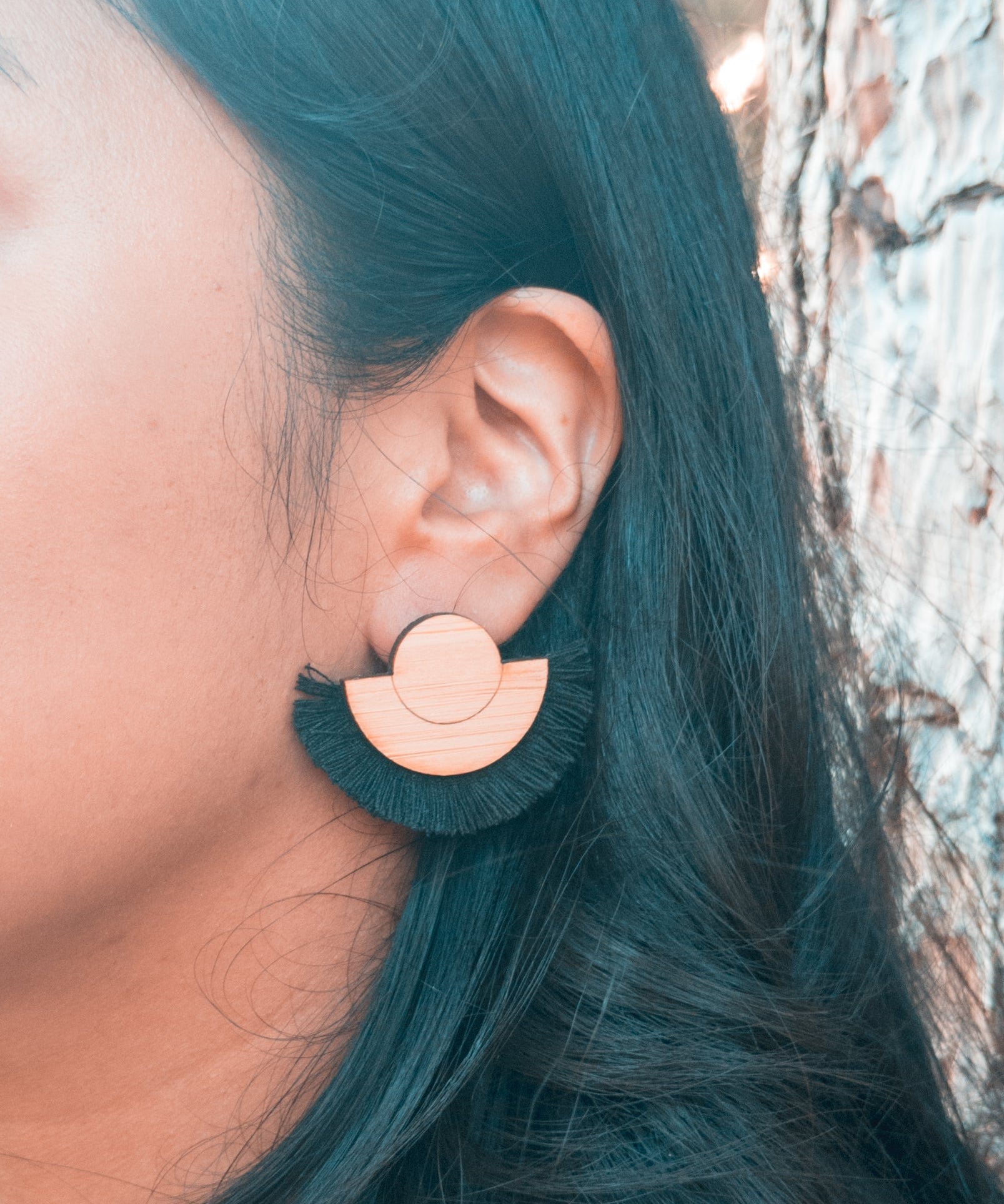 Person wearing a black fringe and wood earring with a tree in the background