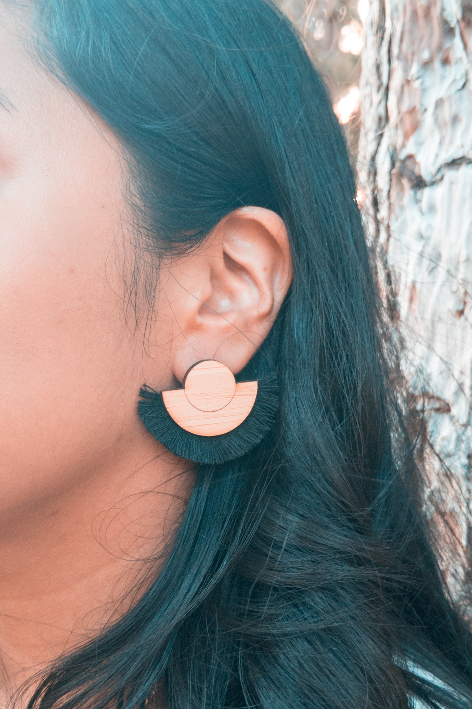 Person wearing a black fringe and wood earring with a tree in the background