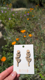 Card with wooden poppy dangles earrings held in front of a natural background with California poppies