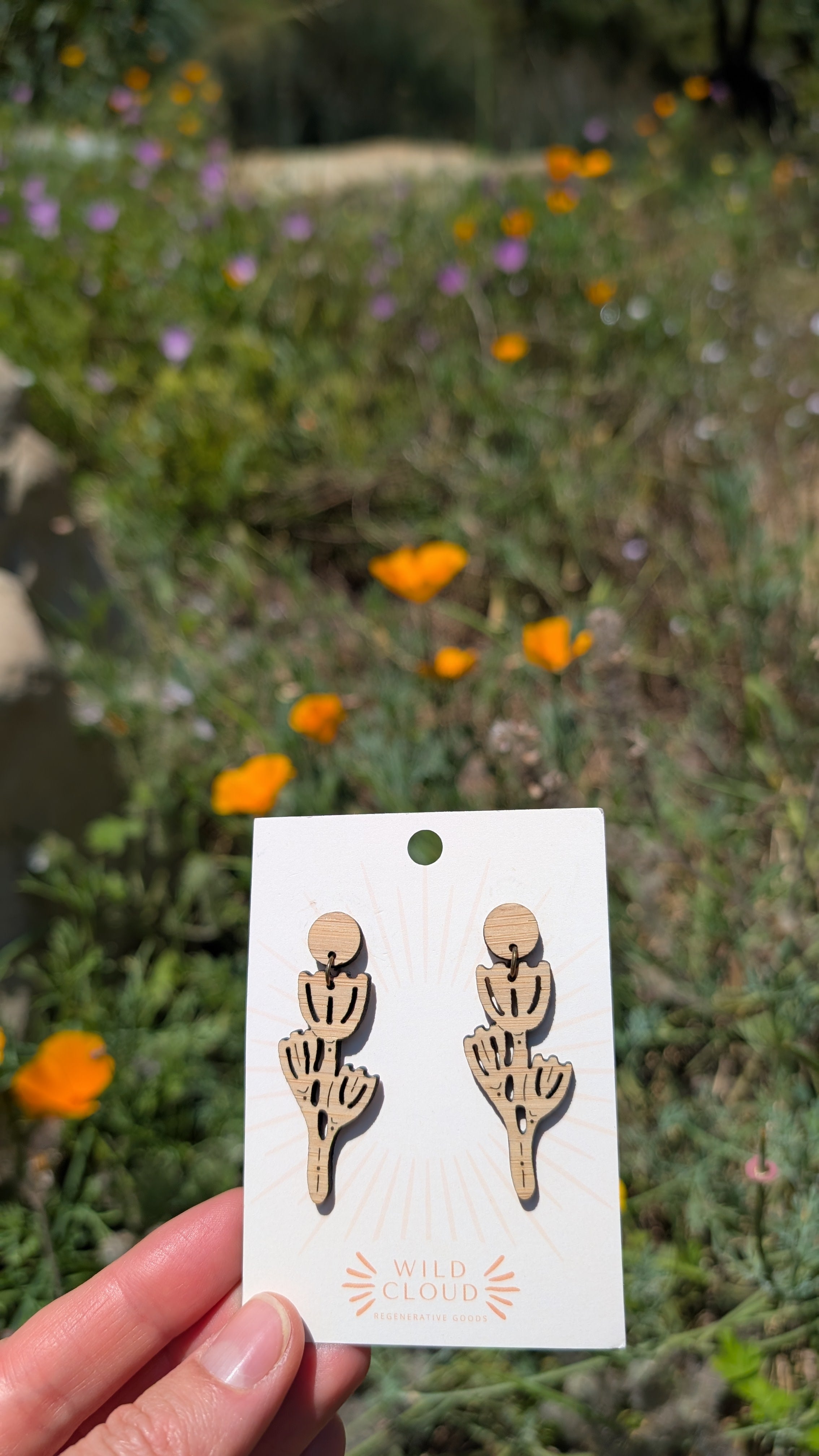 Card with wooden poppy dangles earrings held in front of a natural background with California poppies